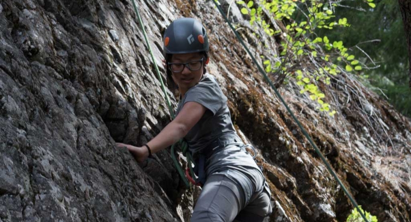 an outward bound student wearing safety gear looks down as they scale a rock wall